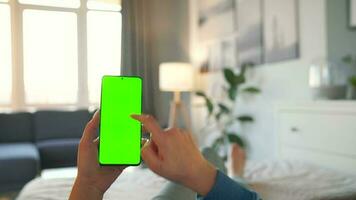 Woman at home lying on a bed and using smartphone with green mock-up screen in vertical mode. video