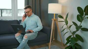 Man in glasses sits on the couch with a laptop and talks on a smartphone. Remote work outside the office video