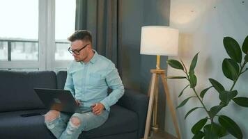 Casual dressed man with glasses sitting on the couch and using laptop to make a video call from home in a modern apartment