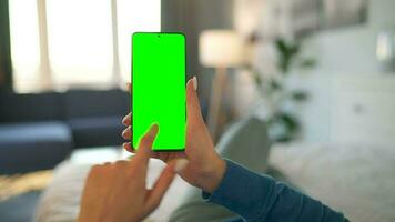 Woman at home lying on a bed and using smartphone with green mock-up screen in vertical mode. video