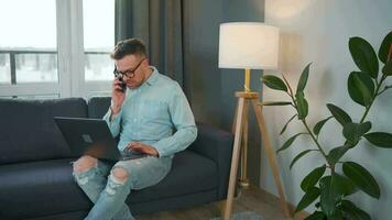 Man in glasses sits on the couch with a laptop and talks on a smartphone. Remote work outside the office video