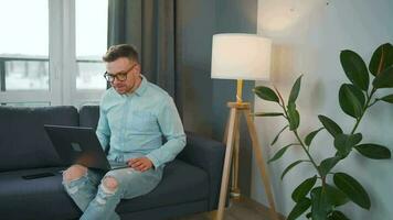 Casual dressed man with glasses sitting on the couch and using laptop to make a video call from home in a modern apartment