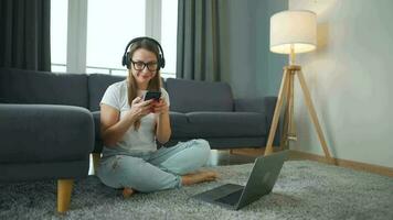 mine de rien habillé femme avec écouteurs est séance sur tapis avec portable et téléphone intelligent dans confortable chambre. elle communique et travaux à distance. éloigné travail à l'extérieur le bureau. video