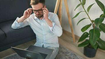 barbu homme avec des lunettes séance sur tapis avec portable et travail dans confortable chambre. éloigné travail à l'extérieur le bureau. video