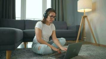 beiläufig gekleidet Frau mit Kopfhörer ist Sitzung auf Teppich mit Laptop im gemütlich Zimmer. sie kommuniziert und funktioniert entfernt. Fernbedienung Arbeit draußen das Büro. video