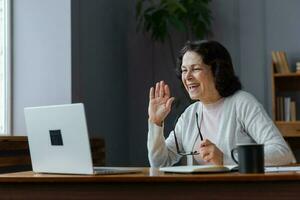 Happy mature blonde lady sitting on couch, holding video call with friends, head shot close up portrait. Web cam view laughing middle aged woman having fun, talking with grown up children online. photo