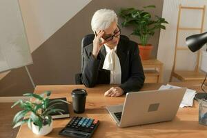 Senior woman touching temples experiencing stress in office. Mature lady tired of working feeling headache sick rubbing temples forehead. Long laptop usage computer vision syndrome. Office syndrome. photo