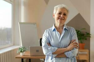 Portrait of confident stylish european middle aged senior woman at workplace. Stylish older mature 60s gray haired lady businesswoman in modern office. Boss leader teacher professional worker. photo