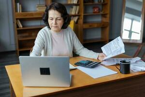 Middle aged senior woman sit with laptop and paper document, Pensive older mature lady reading paper bill pay online at home managing bank finances calculating taxes planning loan debt pension payment photo