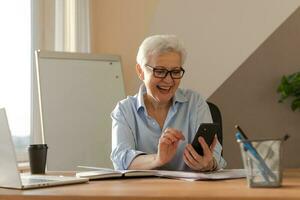 Confident stylish european middle aged senior woman using smartphone at workplace. Stylish older mature 60s gray haired lady businesswoman with cell phone in office. Boss leader using internet apps. photo