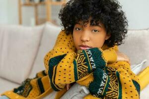 Beautiful african american girl with afro hairstyle sitting on sofa at home indoor. Young african woman with curly hair in living room. Beautiful people natural beauty concept. photo