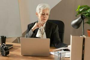 Confident stylish european middle aged senior woman reading financial report documentation statistics. Stylish older mature 60s gray haired lady businesswoman in office. Boss leader teacher. photo