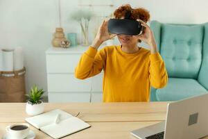 African american young woman wearing using virtual reality metaverse VR glasses headset at home. Girl puts on virtual reality helmet. Technology simulation hi-tech videogame concept. photo