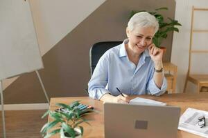 Confident stylish european middle aged senior woman using laptop at workplace. Stylish older mature 60s gray haired lady businesswoman sitting at office table. Boss leader teacher professional worker. photo