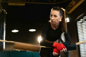 mujer combatiente niña fuerza. mujer combatiente con rojo boxeo envuelve protector vendajes en pie en boxeo anillo propensión en cuerdas esperando descansando fuerte poderoso muchacha. fuerza ajuste cuerpo rutina de ejercicio capacitación. foto