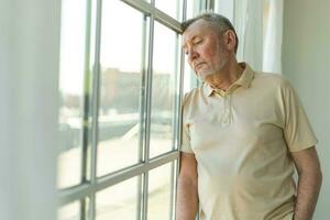 Sad tired lonely disappointed older senior man at home alone. Unhappy grandfather looking sadly outside window experiencing grief relative death bad news. Stressed elder man suffering from loneliness. photo