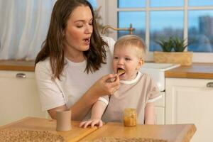 Happy family at home. Mother feeding her baby boy from spoon in kitchen. Little toddler child with messy funny face eats healthy food at home. Young woman mom giving food to kid son. photo