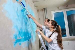 Pareja en nuevo hogar durante reparar trabajos pintura pared juntos. contento familia participación pintar rodillo pintura pared con azul color pintar en nuevo casa. hogar renovación bricolaje renovar hogar concepto. foto