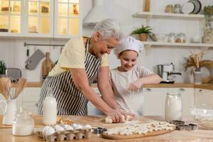 Happy family in kitchen. Grandmother and granddaughter child cook in kitchen together. Grandma teaching kid girl knead dough bake cookies. Household teamwork helping family generations concept. photo