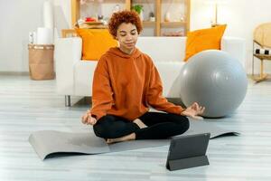 Yoga mindfulness meditation. Young healthy african girl practicing yoga at home. Woman sitting in lotus pose on yoga mat meditating smiling relaxing indoor. Girl doing breathing practice, yoga at home photo