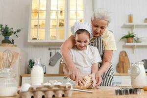 contento familia en cocina. abuela y nieta niño cocinar en cocina juntos. abuela enseñando niño niña amasar masa hornear galletas. casa trabajo en equipo Ayudar familia generaciones concepto. foto