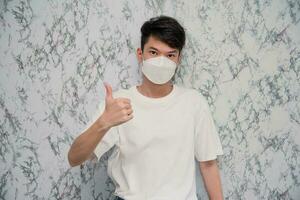 Young Man puts on a face mask showing thumbs up and looking serious isolated on White background,protecting from virus during quarantine.Covid-19 photo