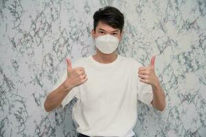 Young Man puts on a face mask showing thumbs up and looking serious isolated on White background,protecting from virus during quarantine.Covid-19 photo