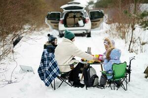 joven mujer con niños en invierno bosque en un picnic. madre y Tres niños en contra ellos coche con abierto trompa. foto