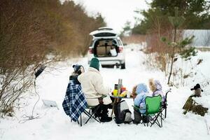 joven mujer con niños en invierno bosque en un picnic. madre y Tres niños en contra ellos coche con abierto trompa. foto