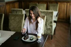 Young smiling brunette woman sit in coffee shop cafe restaurant indoors and eat chocolate brownie dessert cake. photo