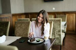 Young smiling brunette woman sit in coffee shop cafe restaurant indoors and eat chocolate brownie dessert cake. photo