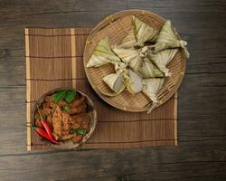 Traditional dry beef curry rendang in coconut shell with diamond shaped woven palm leaf pouch steamed glutenous rice cake ketupat kupat tipat on bamboo tray mat rustic wooden table background photo