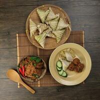 Traditional dry beef curry rendang in coconut shell with diamond shaped woven palm leaf pouch steamed glutenous rice cake ketupat kupat tipat on bamboo tray mat rustic wooden table background photo