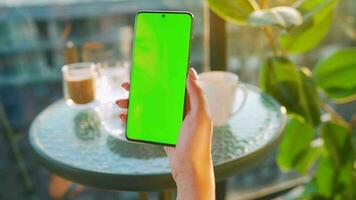 Woman sitting in a cafe and using smartphone with green mock-up screen in vertical mode. Girl browsing Internet video