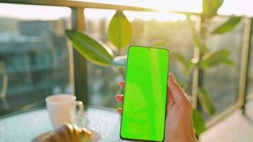 Woman sitting in a cafe and using smartphone with green mock-up screen in vertical mode. Girl browsing Internet video