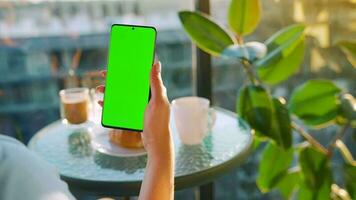 Woman sitting in a cafe and using smartphone with green mock-up screen in vertical mode. Girl browsing Internet video