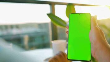 Woman sitting in a cafe and using smartphone with green mock-up screen in vertical mode. Girl browsing Internet video