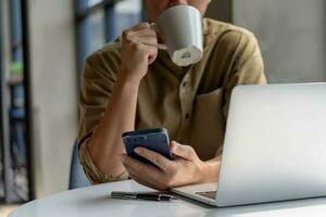 man asian drinking coffee and looking mobile phone computer laptop and at desk. Business technology digital checking emails or browsing social media while taking a quick coffee break. photo