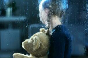 Girl with Teddy Bear Behind Window Covered by Rain Drops photo