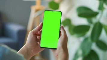 Woman at home using smartphone with green mock-up screen in vertical mode. Girl browsing Internet, watching content video