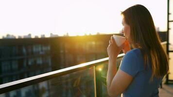 kvinna med en kopp av kaffe stående på de balkong och beundra de solnedgång video