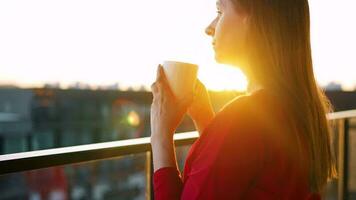 mulher com uma copo do café em pé em a sacada e admirar a pôr do sol video