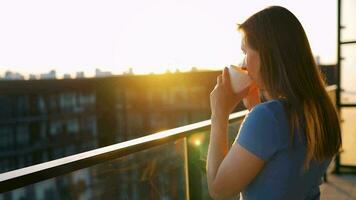 mulher com uma copo do café em pé em a sacada e admirar a pôr do sol video