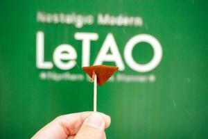 Otaru, Hokkaido, Japan, 2018 - Hand holding small wood stick with a free chocolate on LeTAO sign cafe background. LeTAO is the popular sweet cafe of tourist when come to visit Otaru city. photo