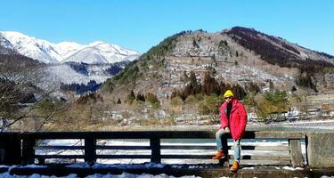 Tourist man in red coat and yellow knit hat sits on bridge rail with tree, snow on mountain and clear blue sky background with copy space at Japan. People travel in Asia with natural and landmarks. photo