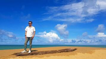 Asian man in white shirt and jeans, wearing sunglasses, stand on timber with sea, blue sky and cloud background with copy space at Mai Khao beach Phuket, Thailand. Tourist, Relax, landmark, travel. photo