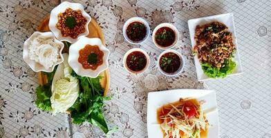 Top view of rice noodles with fresh vegetable and sweet sauce in wooden tray with spicy sauce, papaya salad Thai style and Spicy minced pork on table for lunch at Thai local restaurant - Asian food photo