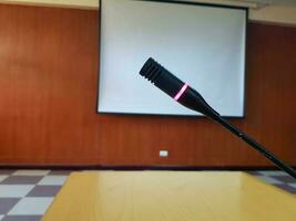 negro micrófono con rojo ligero brillante cuando empujar botón en de madera escritorio para hablando o presentación en conferencia o reunión habitación con blanco proyector tablero borroso antecedentes. dispositivo y herramienta foto