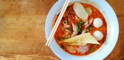 Top view hot spicy noodle with soup, chopsticks, deep fried won-ton meat, fish or chicken ball, crispy pork and in light blue bowl on wooden table or background with left copy space. Asian food photo