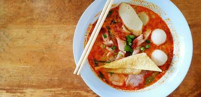 Top view hot spicy noodle with soup, chopsticks, deep fried won-ton meat, fish or chicken ball, crispy pork and in light blue bowl on wooden table or background with left copy space. Asian food photo